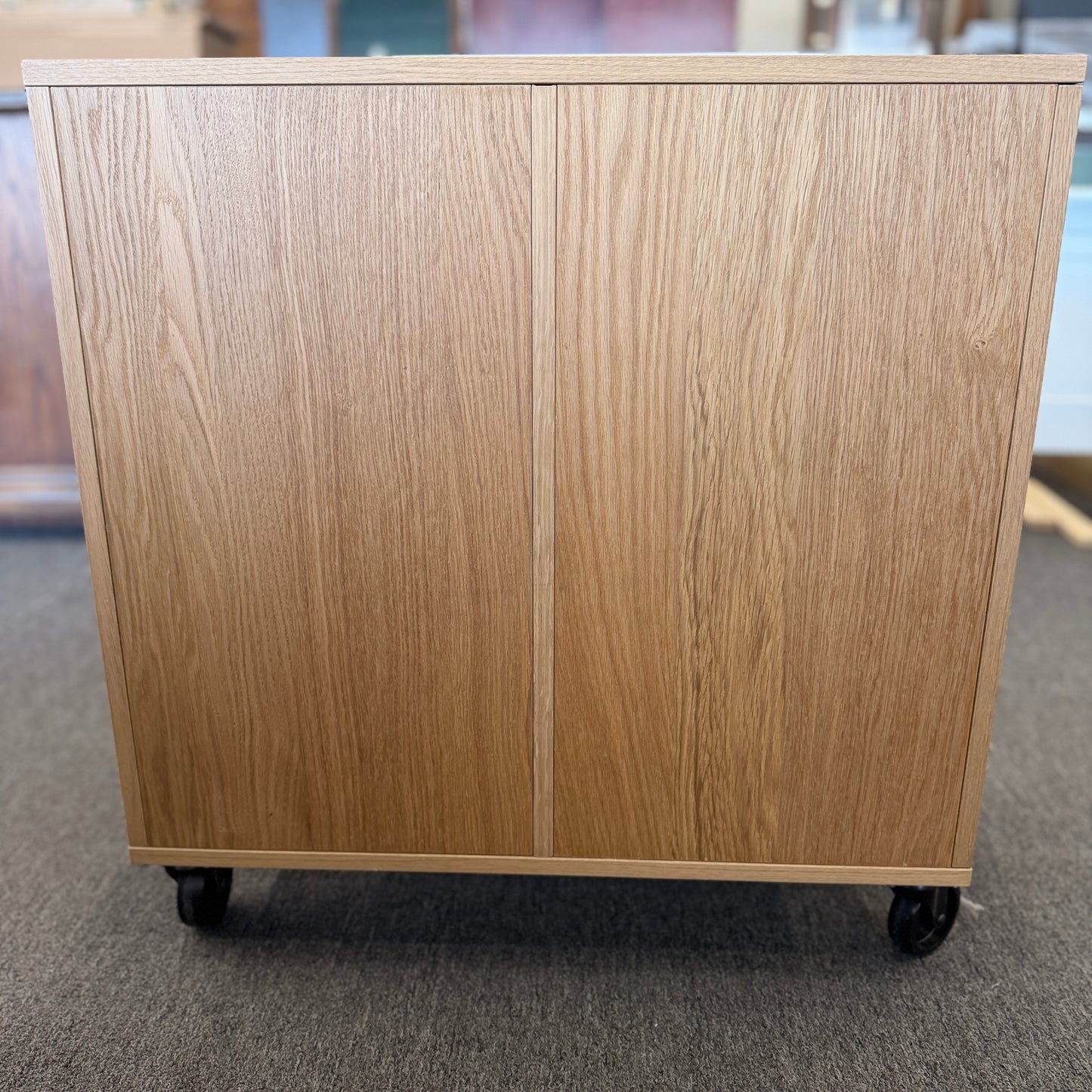 A modern mobile office storage cart with a light oak finish, featuring asymmetrical divided cubby compartments and four black rolling casters.
