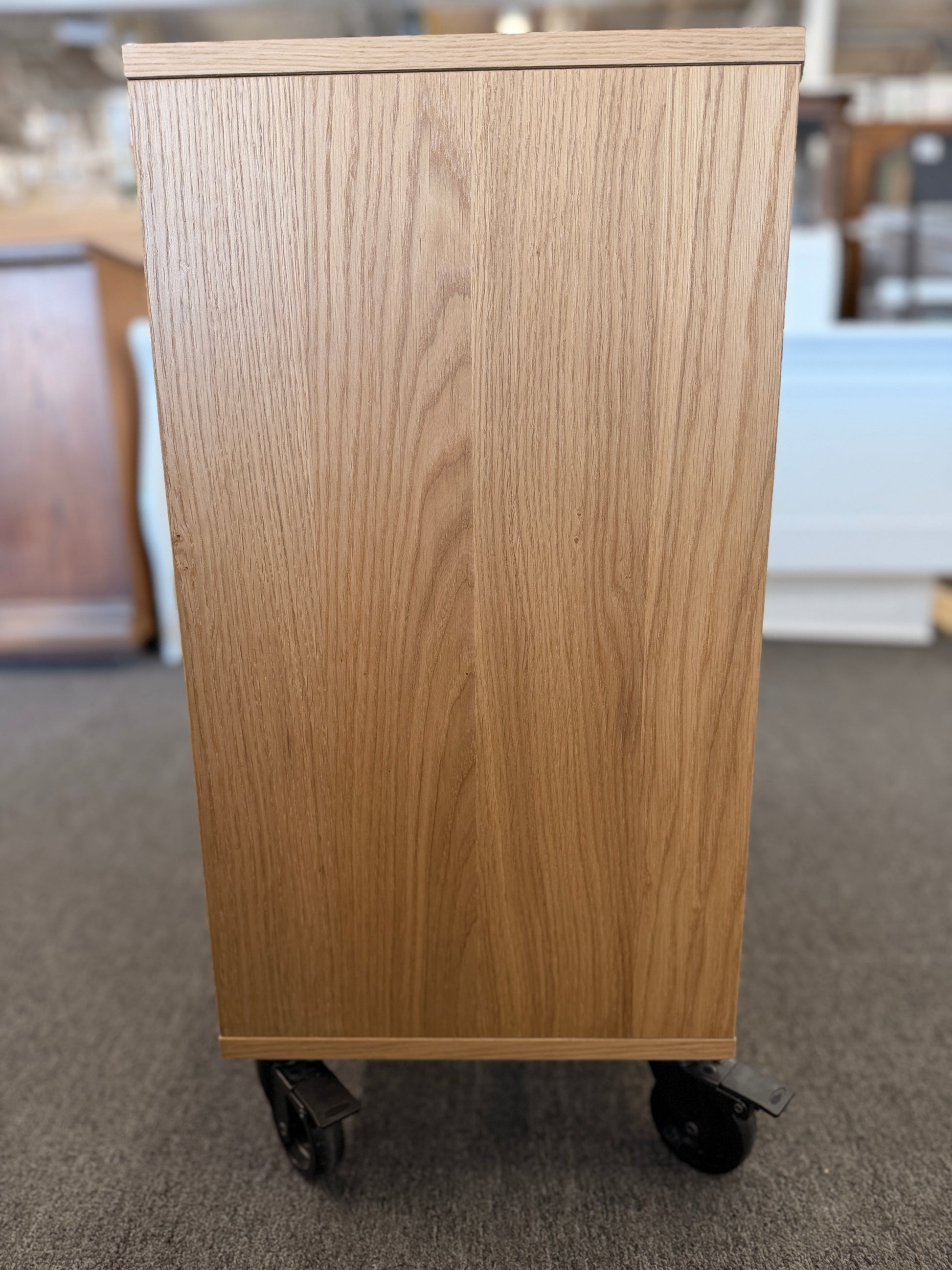 A modern mobile office storage cart with a light oak finish, featuring asymmetrical divided cubby compartments and four black rolling casters.