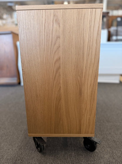 A modern mobile office storage cart with a light oak finish, featuring asymmetrical divided cubby compartments and four black rolling casters.