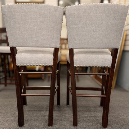 A pair of upholstered high-back counter stools featuring light grey textured fabric, classic metal nailhead trim, and dark-stained wooden legs with footrests.
