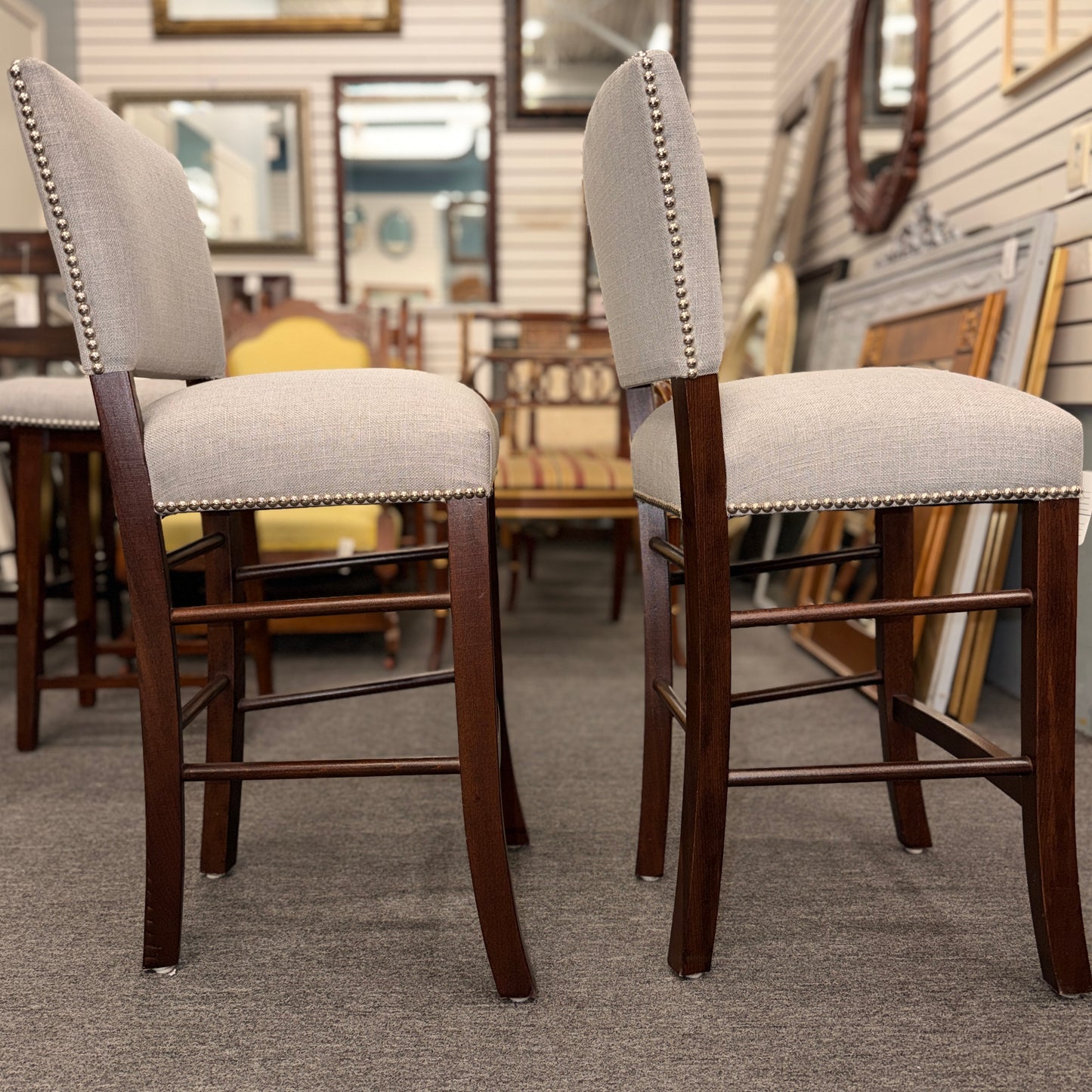 A pair of upholstered high-back counter stools featuring light grey textured fabric, classic metal nailhead trim, and dark-stained wooden legs with footrests.