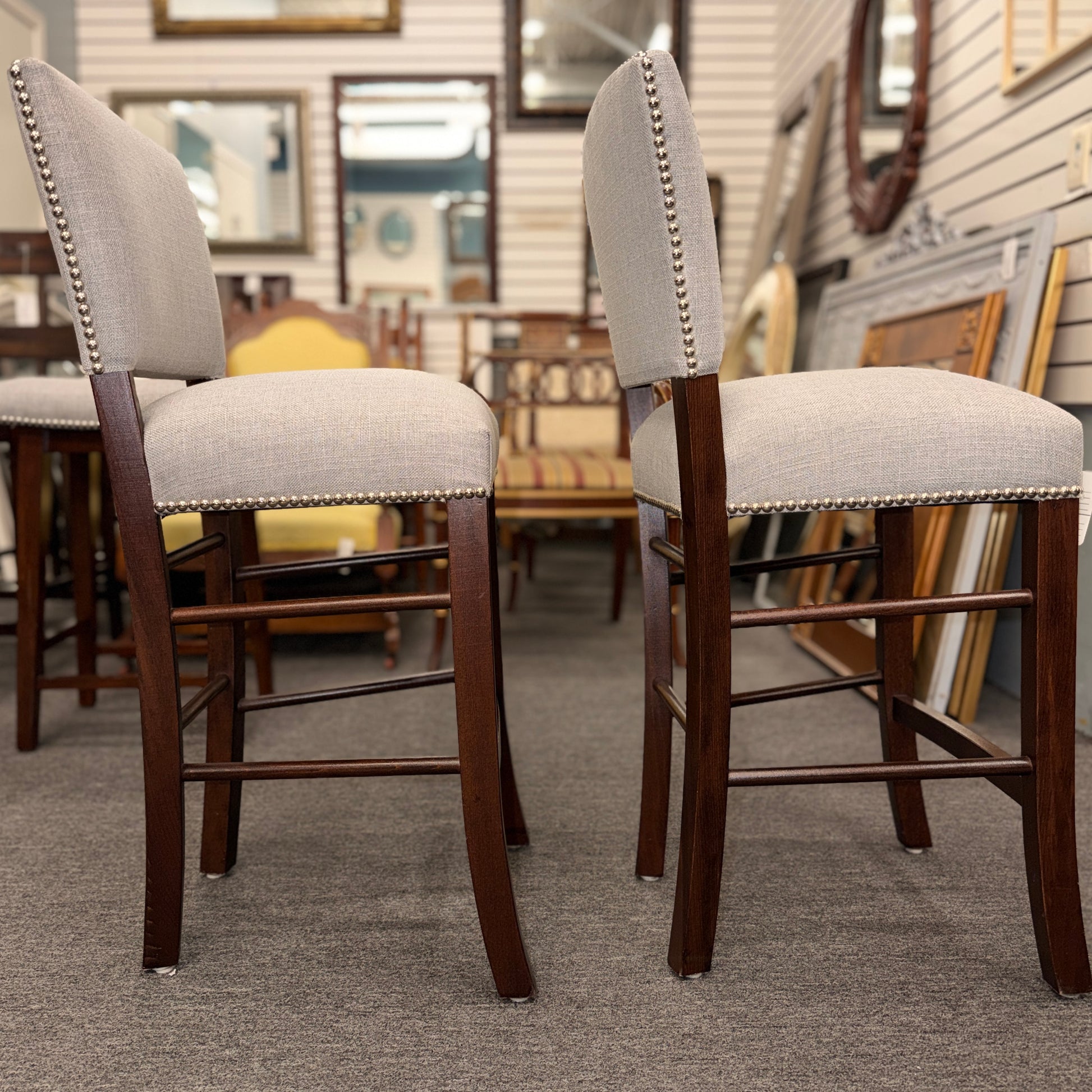 A pair of upholstered high-back counter stools featuring light grey textured fabric, classic metal nailhead trim, and dark-stained wooden legs with footrests.