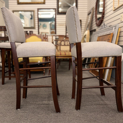 A pair of upholstered high-back counter stools featuring light grey textured fabric, classic metal nailhead trim, and dark-stained wooden legs with footrests.