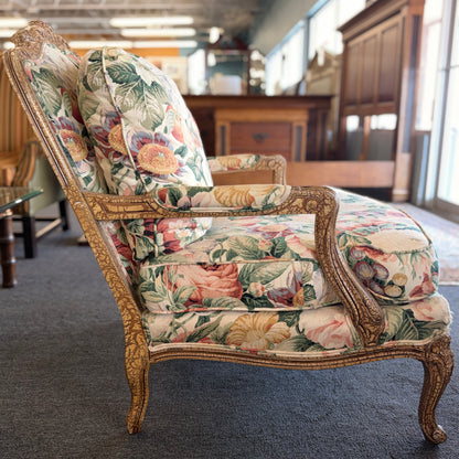A French provincial-style armchair with an intricately carved exposed wooden frame, elegant cabriole legs, and vibrant multicolored floral fabric upholstery.