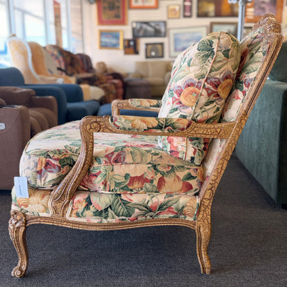 A French provincial-style armchair with an intricately carved exposed wooden frame, elegant cabriole legs, and vibrant multicolored floral fabric upholstery.