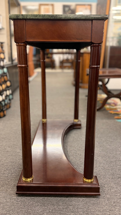 Traditional console table with a rectangular green marble top, dark wood frame, fluted cylindrical legs, gold-toned brass rosette accents, and a solid wood base plinth.