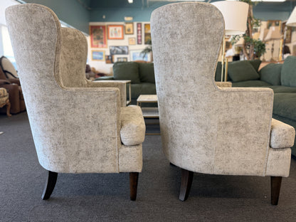 Matching pair of transitional wingback accent armchairs upholstered in textured light beige fabric with dark brown tapered wooden legs.