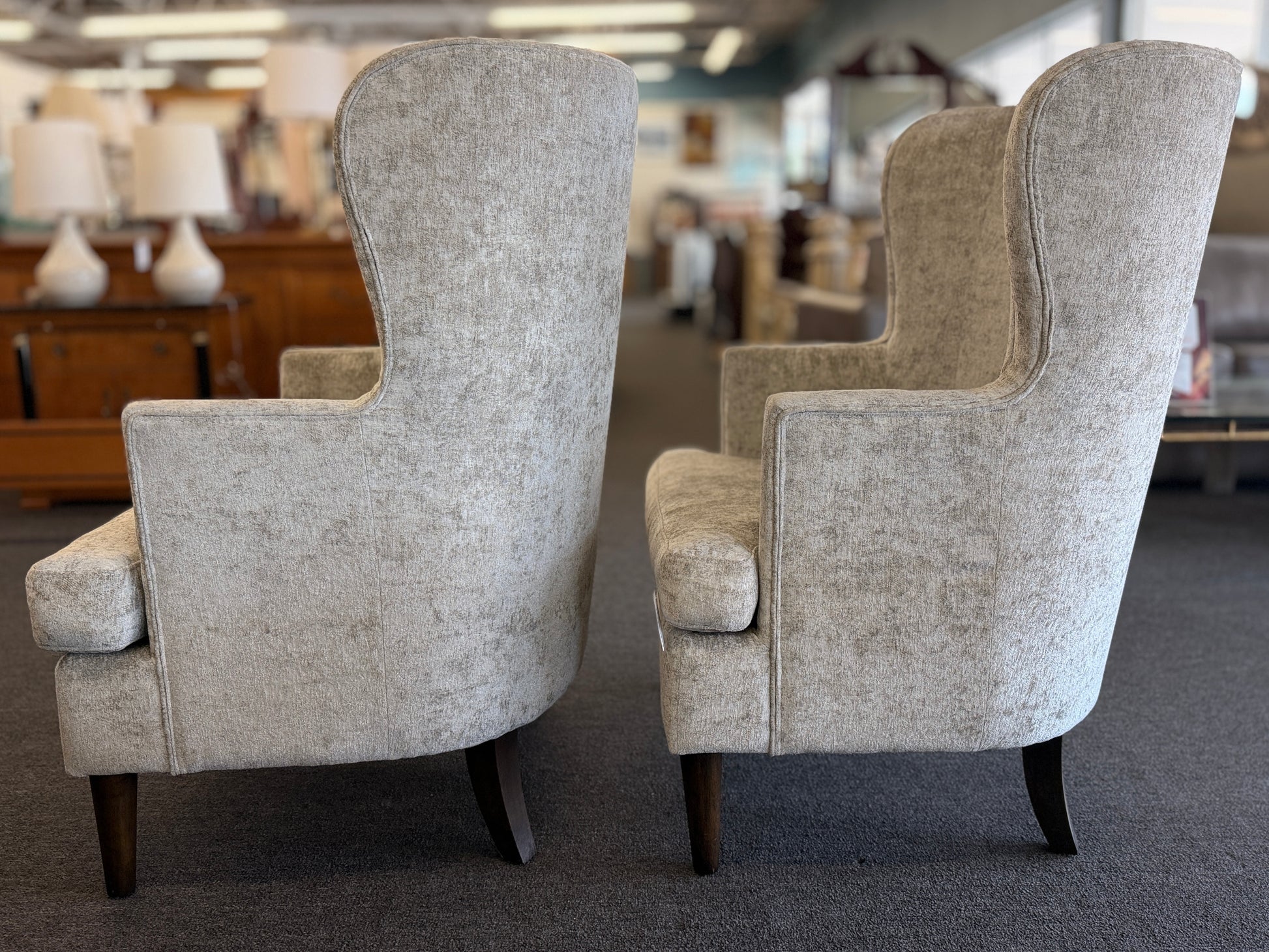 Matching pair of transitional wingback accent armchairs upholstered in textured light beige fabric with dark brown tapered wooden legs.