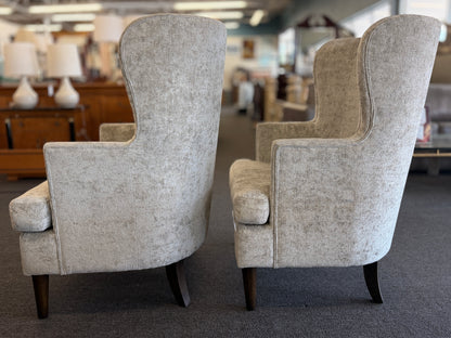 Matching pair of transitional wingback accent armchairs upholstered in textured light beige fabric with dark brown tapered wooden legs.