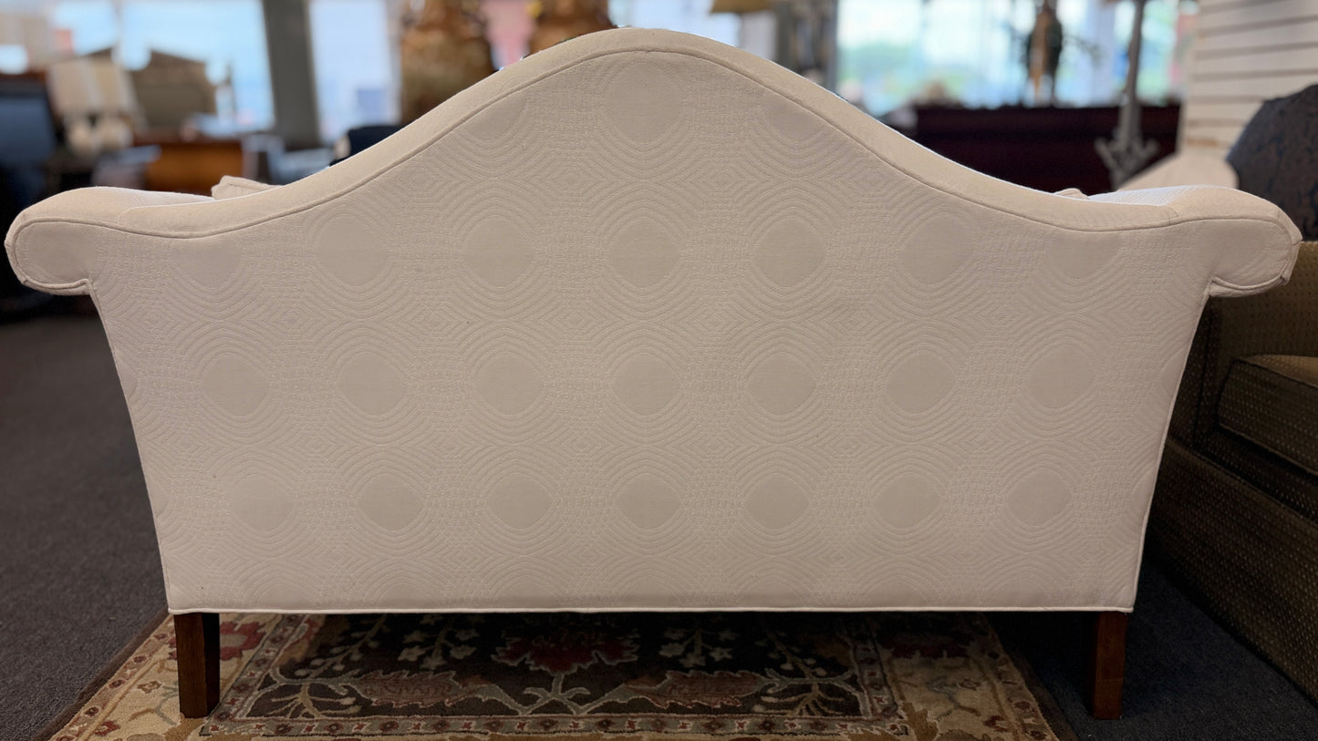 Cream upholstered Ethan Allen camelback loveseat featuring rolled arms, a single seat cushion, two matching throw pillows, and dark tapered wooden legs.
