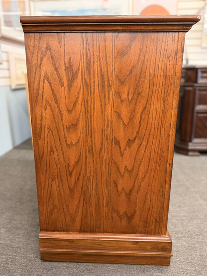 Medium-tone wood storage cabinet featuring three stacked drawers, a glass-paneled cabinet door with interior shelving, and a classic versatile design.