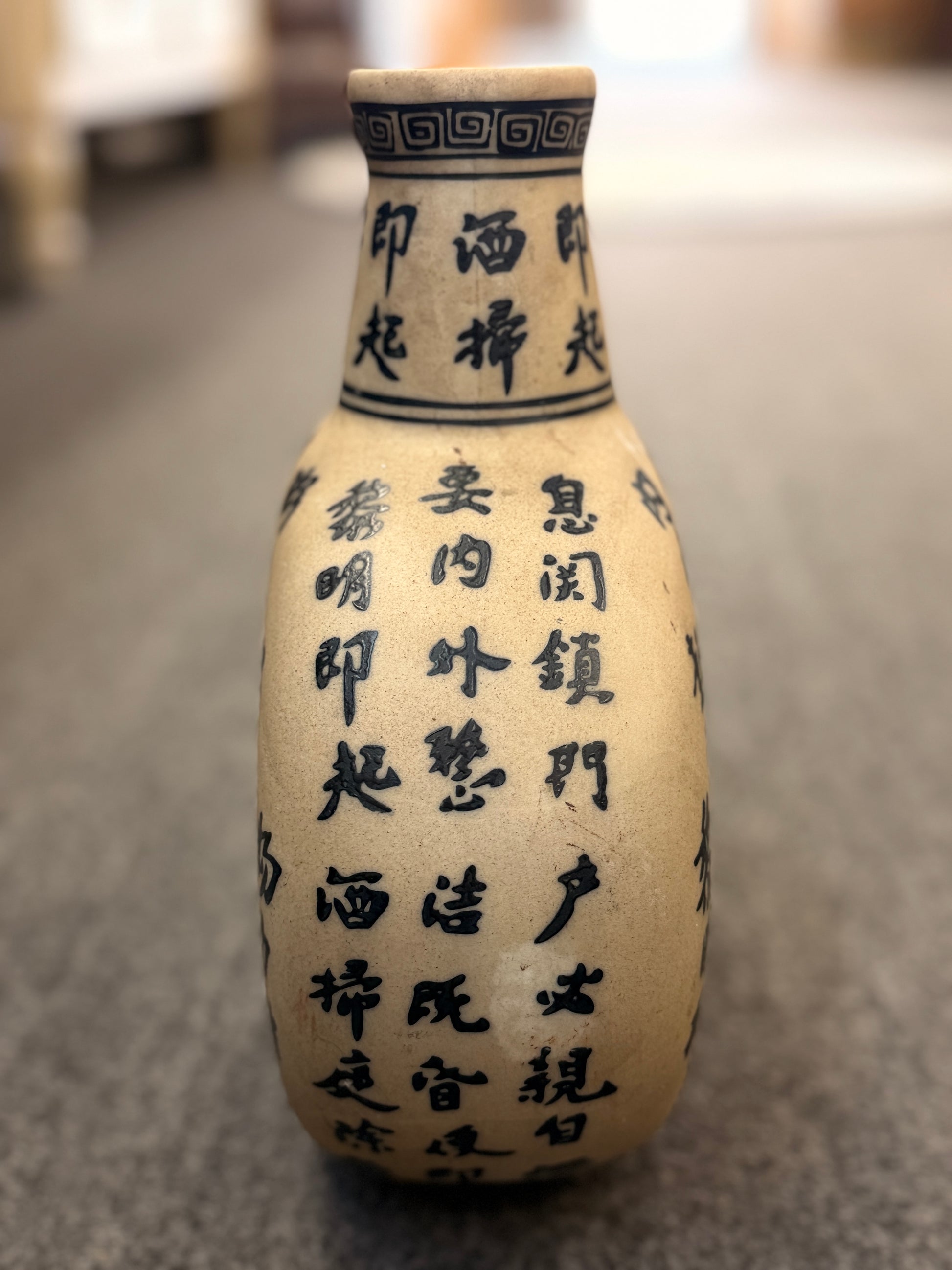 A round, flat-profile ceramic vase featuring black Asian calligraphy painted across a matte sand-colored surface.