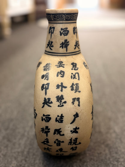 A round, flat-profile ceramic vase featuring black Asian calligraphy painted across a matte sand-colored surface.