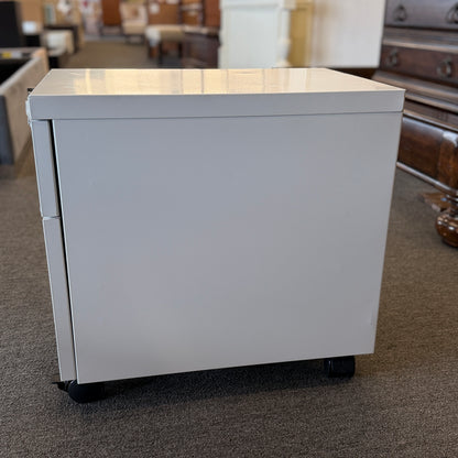 A minimalist two-drawer under-desk pedestal file cabinet featuring curved metal handles, a top lock, and dual casters in good condition.