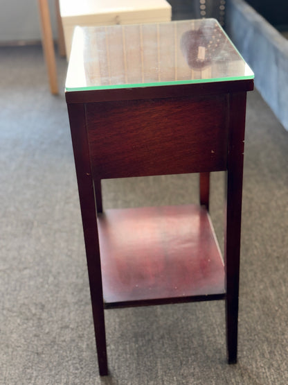 A compact Federal-style wood end table featuring fluted corner carvings, a single drawer with brass bail pull, lower shelf, and a custom glass top in good condition.