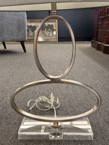 A contemporary table lamp featuring an intertwined dual-ring brushed nickel body, a clear acrylic base, and a textured gray drum shade in excellent condition.