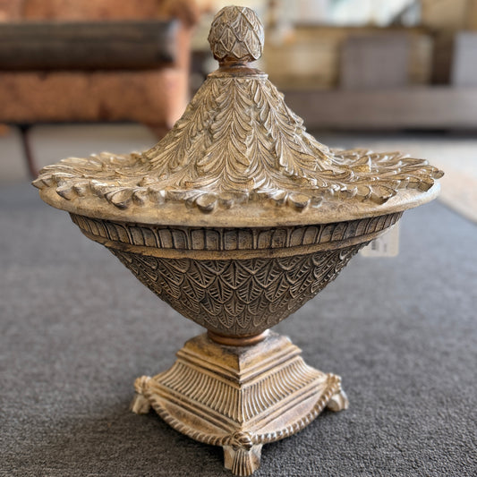A decorative heavily textured ceramic lidded urn covered in overlapping leaf patterns with an antiqued distressed cream and brown finish.