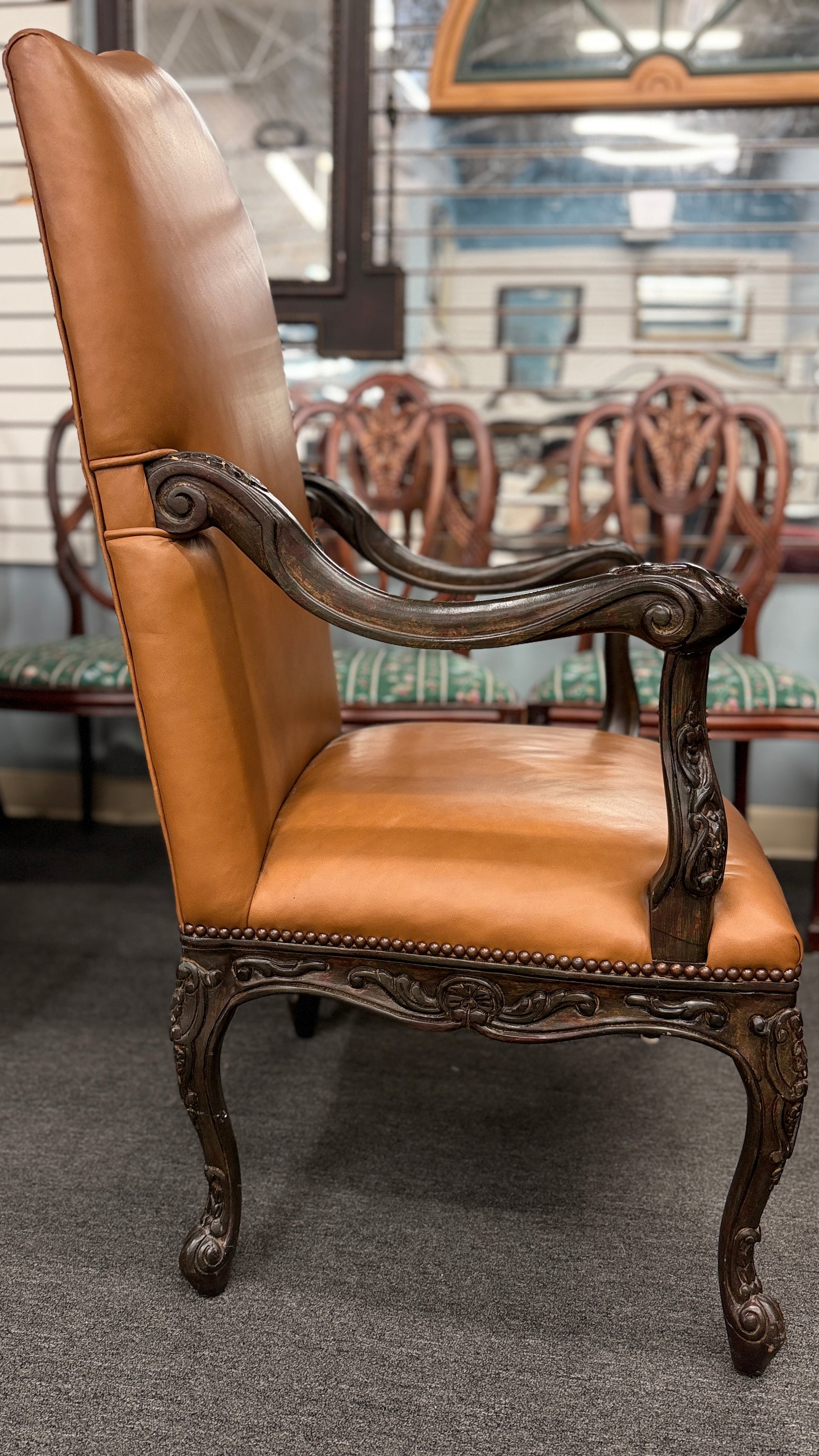 An elegant antique-reproduction accent chair upholstered in smooth tan leather with a hand-carved dark wood frame, cabriole legs, and brass nailhead trim.