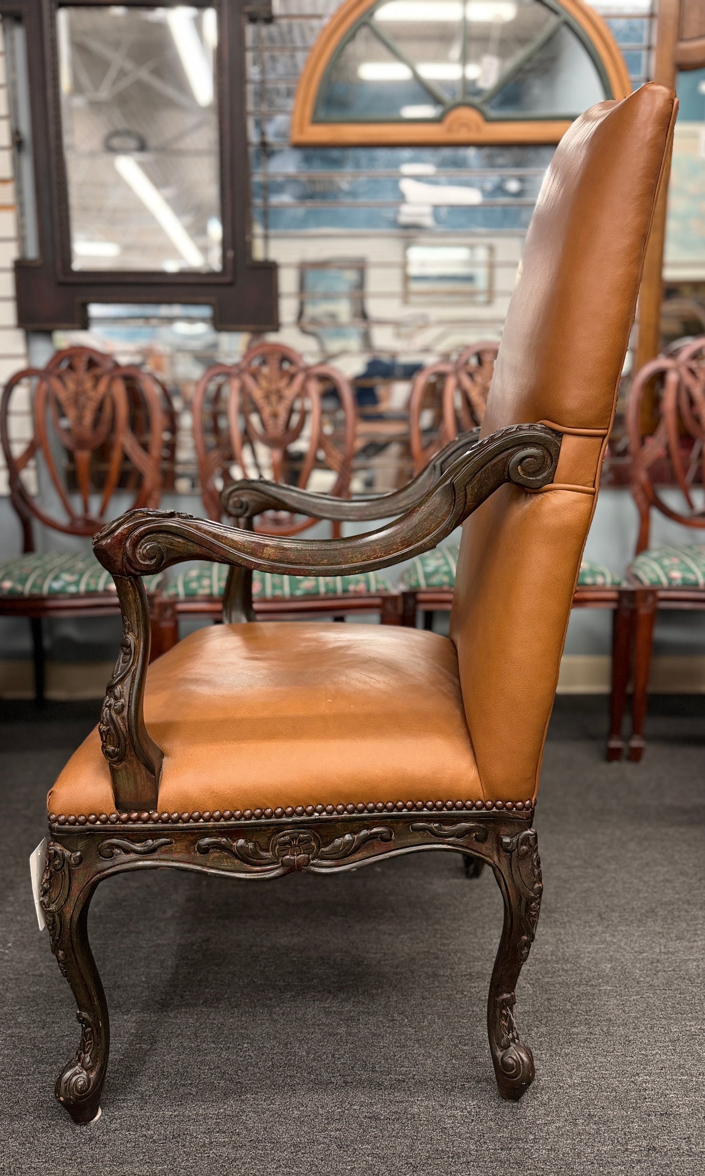 An elegant antique-reproduction accent chair upholstered in smooth tan leather with a hand-carved dark wood frame, cabriole legs, and brass nailhead trim.