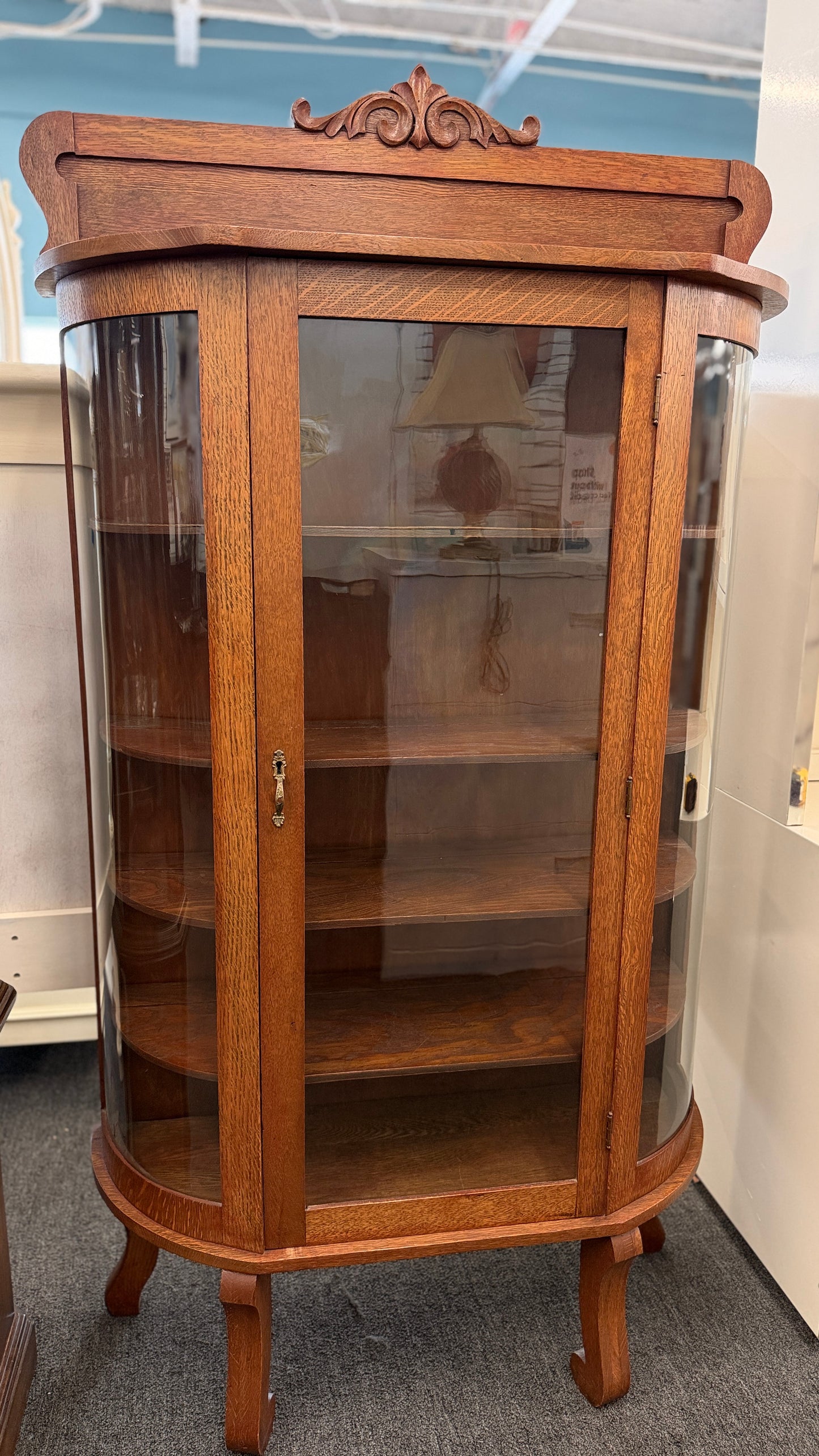 An antique-style oak curio cabinet featuring curved glass sides, an arched door with key hardware, interior shelves, and a carved pediment in good condition.