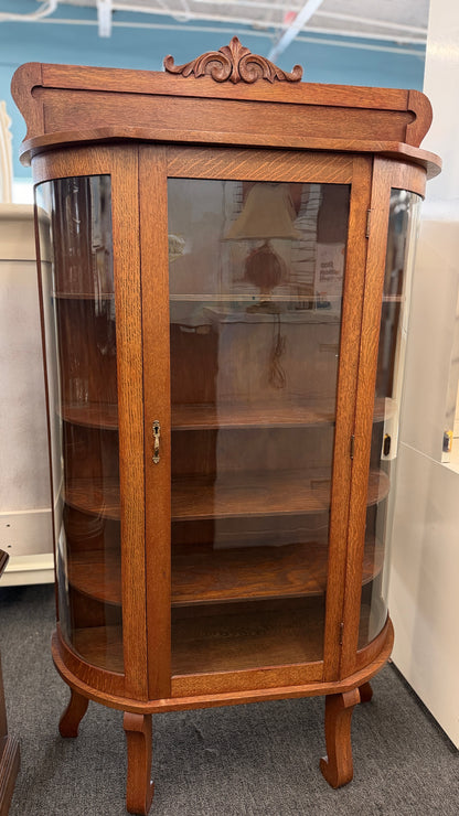 An antique-style oak curio cabinet featuring curved glass sides, an arched door with key hardware, interior shelves, and a carved pediment in good condition.