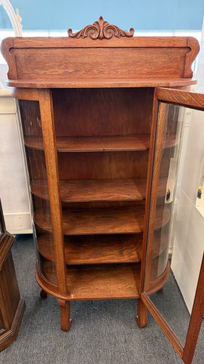 An antique-style oak curio cabinet featuring curved glass sides, an arched door with key hardware, interior shelves, and a carved pediment in good condition.
