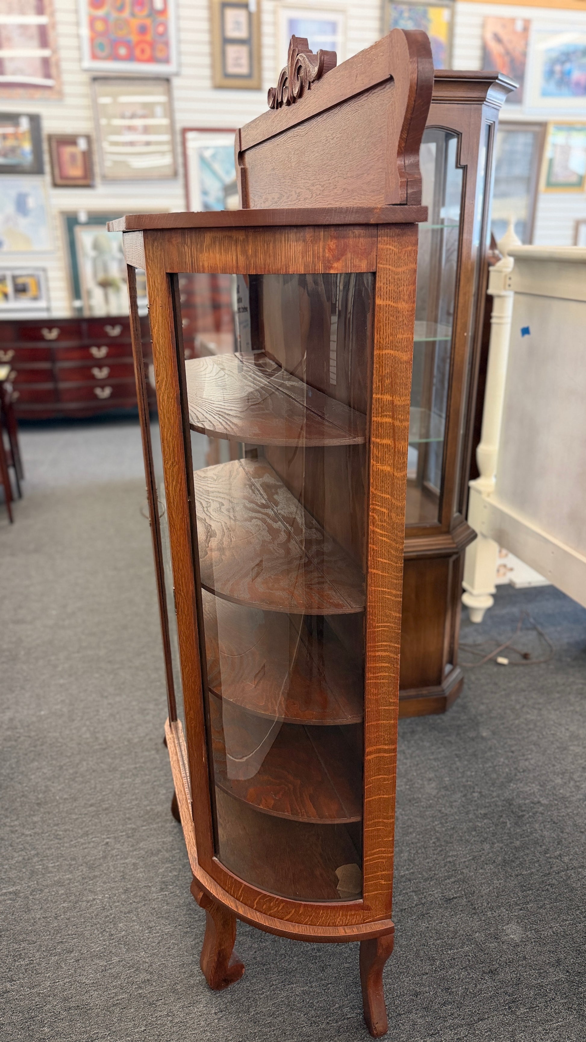An antique-style oak curio cabinet featuring curved glass sides, an arched door with key hardware, interior shelves, and a carved pediment in good condition.