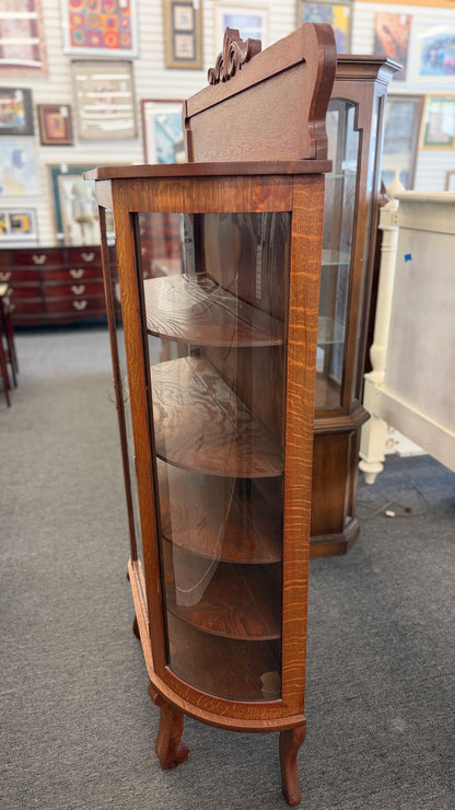 An antique-style oak curio cabinet featuring curved glass sides, an arched door with key hardware, interior shelves, and a carved pediment in good condition.