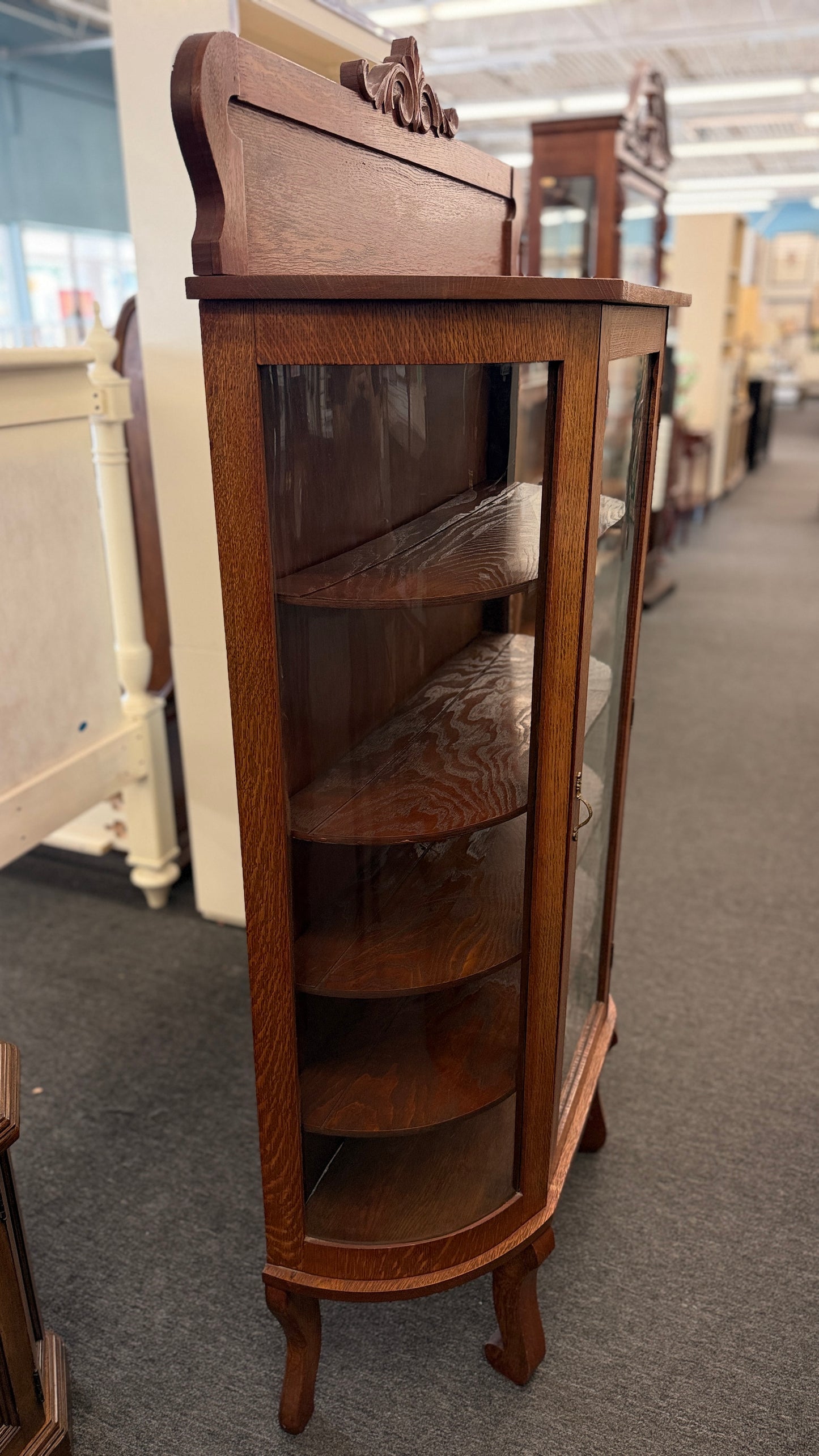 An antique-style oak curio cabinet featuring curved glass sides, an arched door with key hardware, interior shelves, and a carved pediment in good condition.