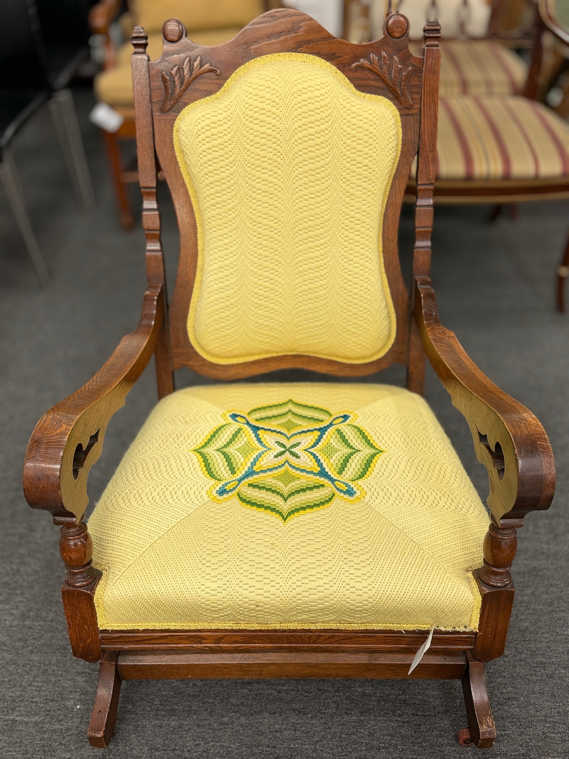 A vintage-style wooden platform rocking chair with an ornate carved crest rail, custom yellow upholstery, and a geometric green and blue embroidered center design.