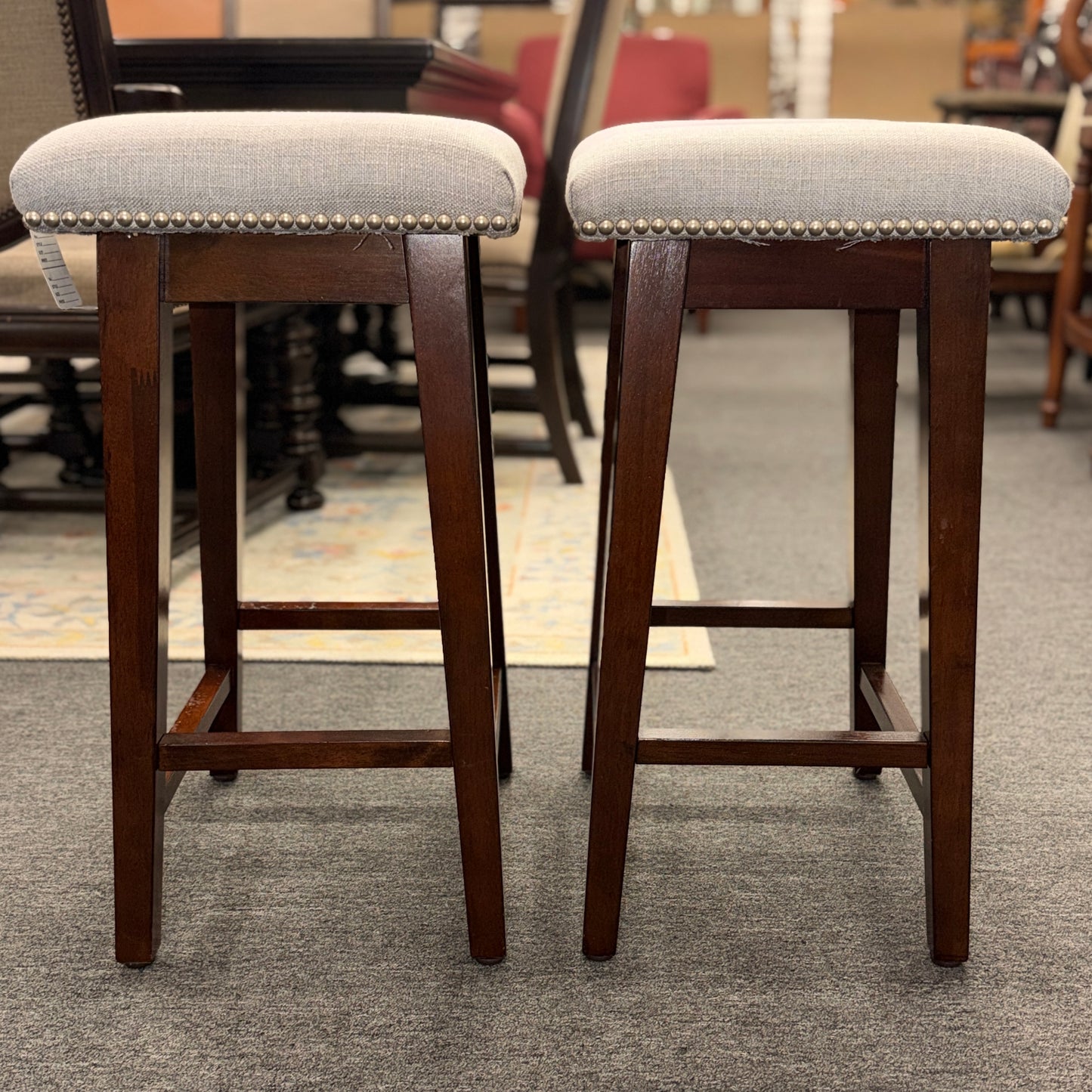 A pair of backless saddle-style counter stools featuring light grey upholstered seats with metal nailhead trim and dark wood bases with built-in footrests.