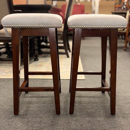 A pair of backless saddle-style counter stools featuring light grey upholstered seats with metal nailhead trim and dark wood bases with built-in footrests.