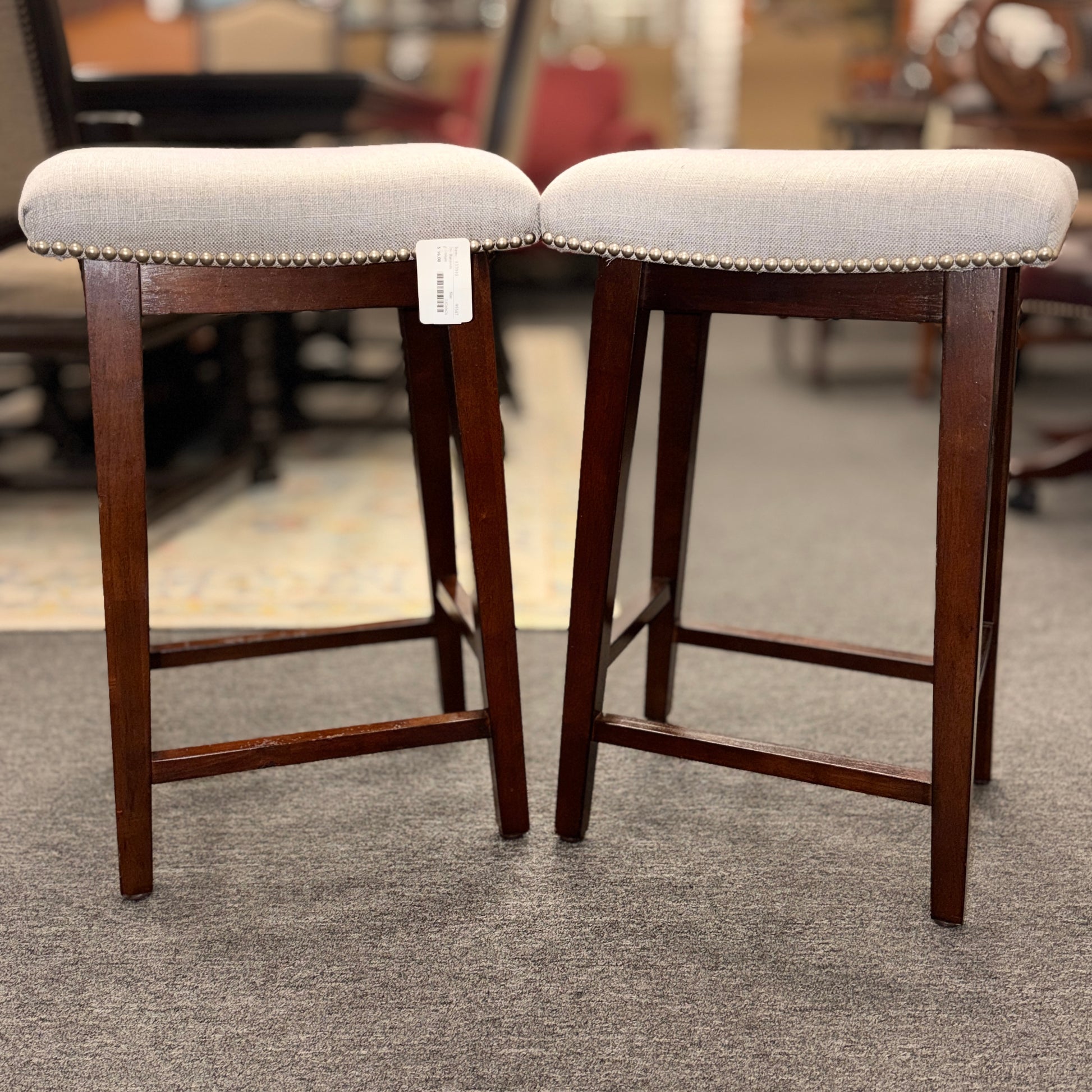 A pair of backless saddle-style counter stools featuring light grey upholstered seats with metal nailhead trim and dark wood bases with built-in footrests.