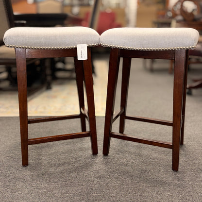 A pair of backless saddle-style counter stools featuring light grey upholstered seats with metal nailhead trim and dark wood bases with built-in footrests.