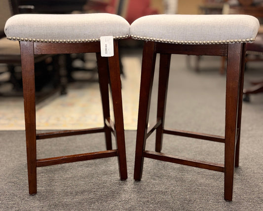 A pair of backless saddle-style counter stools featuring light grey upholstered seats with metal nailhead trim and dark wood bases with built-in footrests.