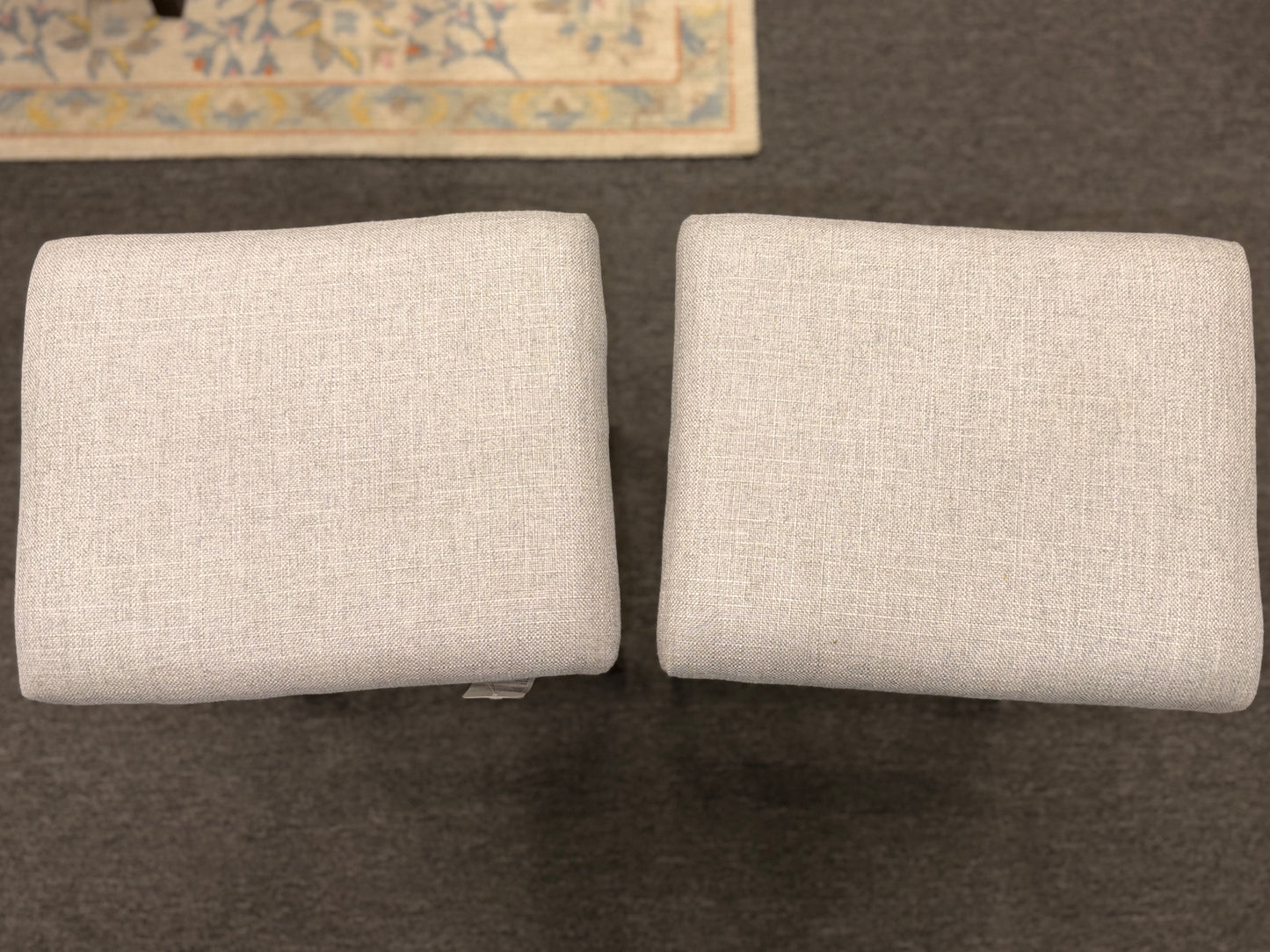 A pair of backless saddle-style counter stools featuring light grey upholstered seats with metal nailhead trim and dark wood bases with built-in footrests.