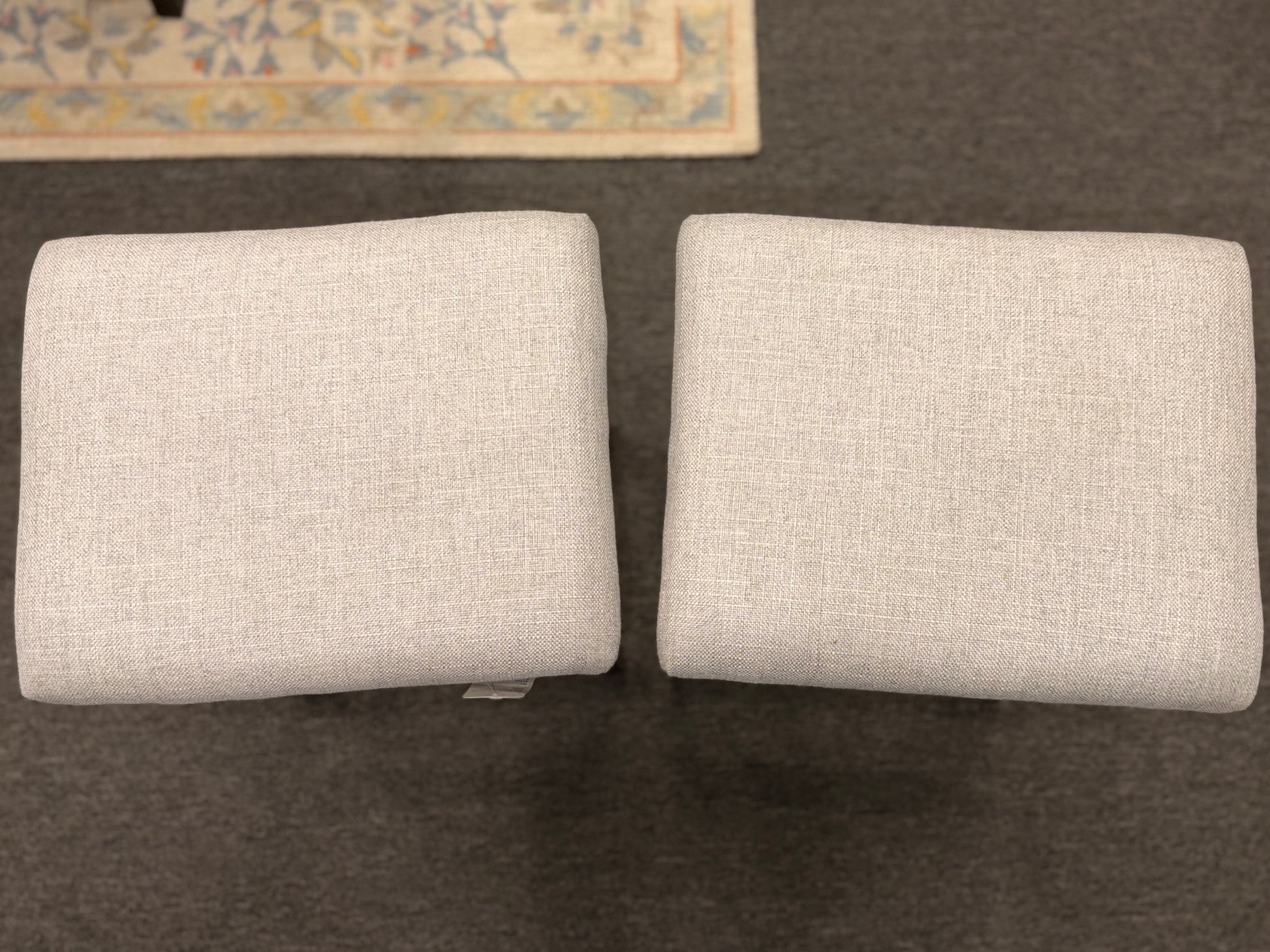 A pair of backless saddle-style counter stools featuring light grey upholstered seats with metal nailhead trim and dark wood bases with built-in footrests.