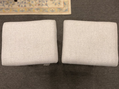 A pair of backless saddle-style counter stools featuring light grey upholstered seats with metal nailhead trim and dark wood bases with built-in footrests.