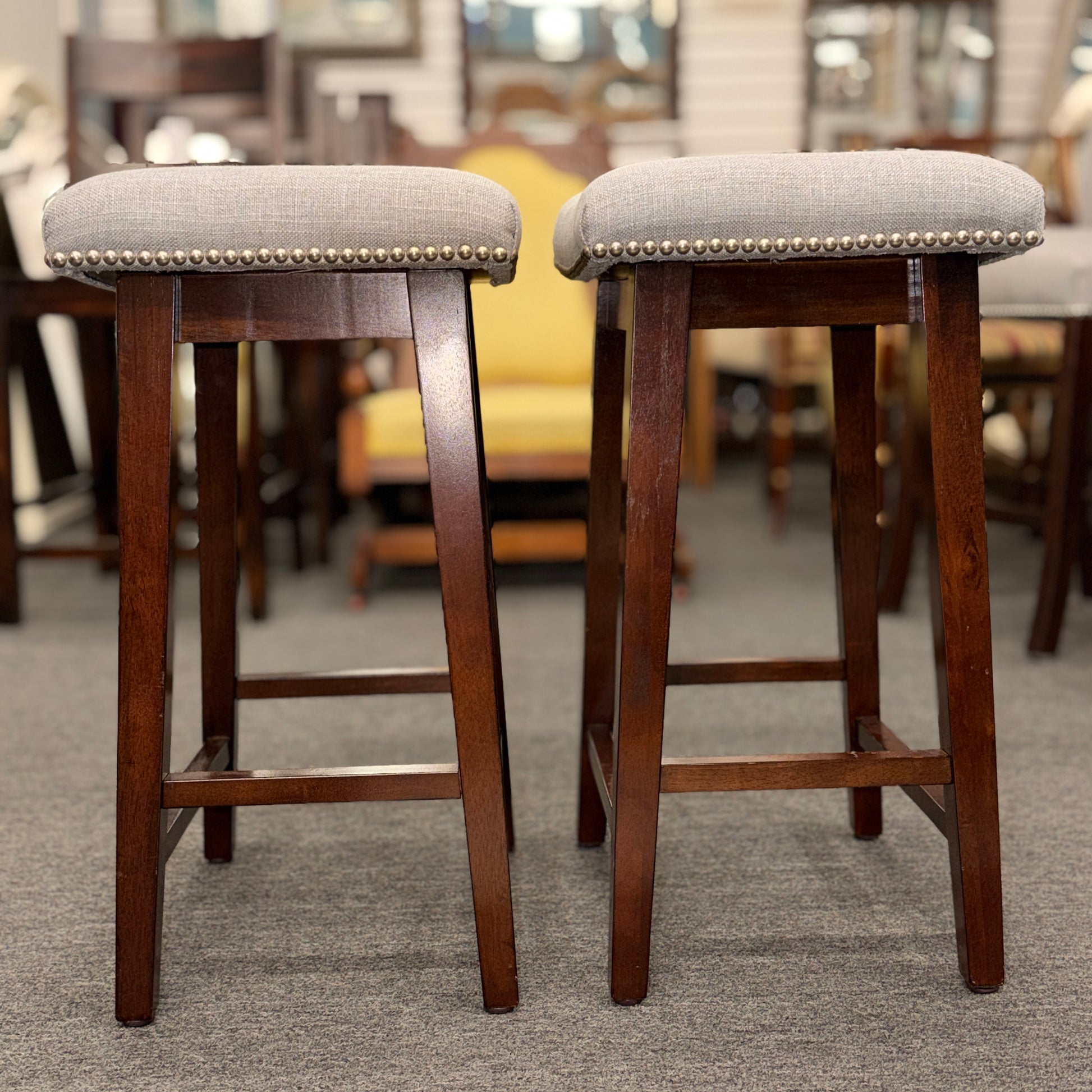 A pair of backless saddle-style counter stools featuring light grey upholstered seats with metal nailhead trim and dark wood bases with built-in footrests.