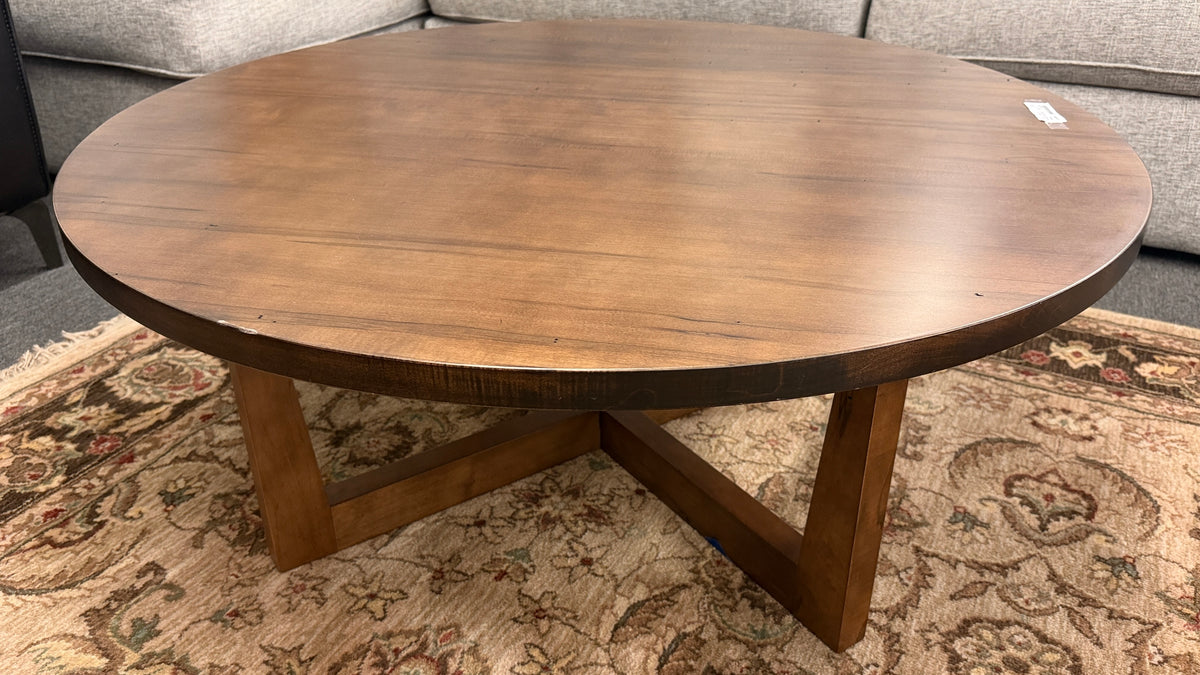 A round rustic-contemporary Bassett wooden coffee table in a warm walnut finish supported by an angular intersecting cross-base in good condition.