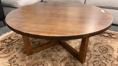 A round rustic-contemporary Bassett wooden coffee table in a warm walnut finish supported by an angular intersecting cross-base in good condition.