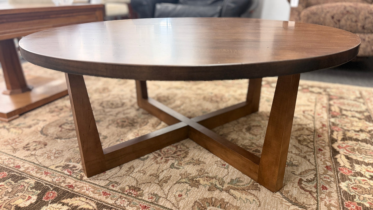 A round rustic-contemporary Bassett wooden coffee table in a warm walnut finish supported by an angular intersecting cross-base in good condition.