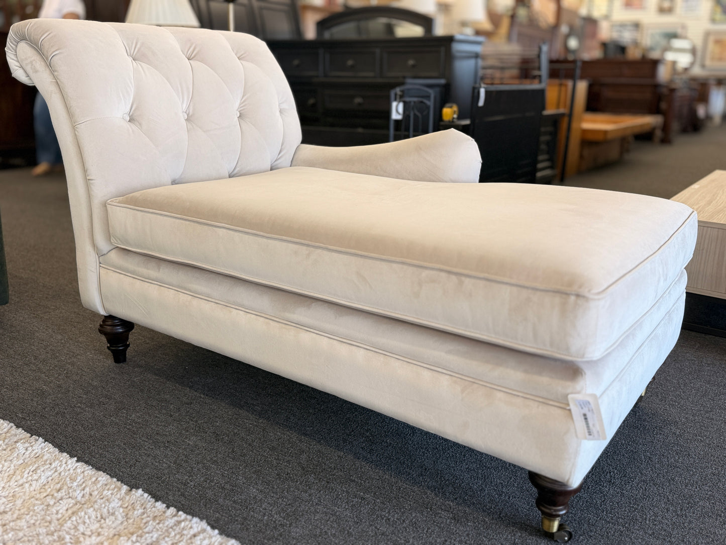 A vintage-style Bassett chaise lounge with a tufted rolled backrest, plush fabric cushion, and turned wooden legs on front casters.