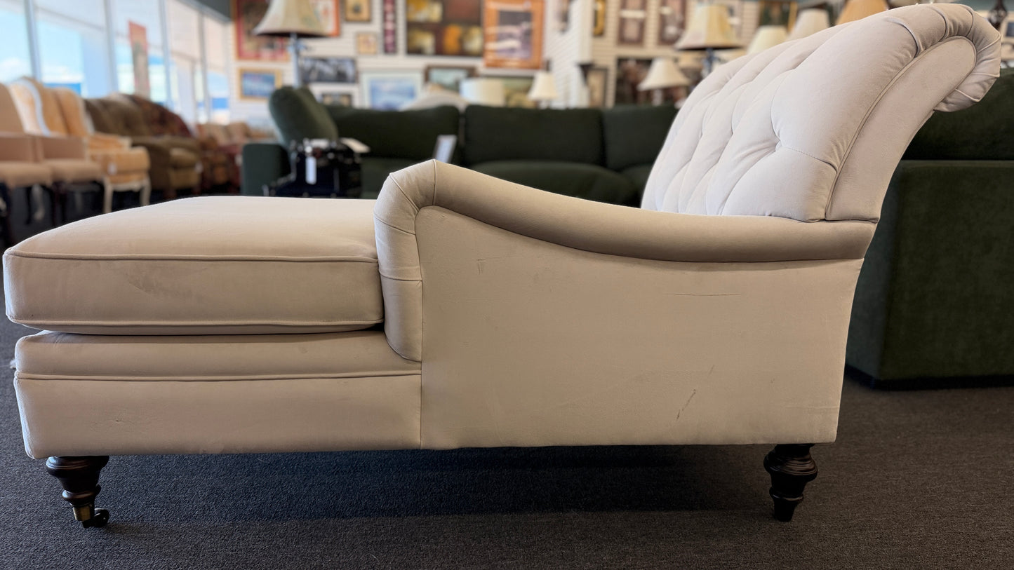 A vintage-style Bassett chaise lounge with a tufted rolled backrest, plush fabric cushion, and turned wooden legs on front casters.