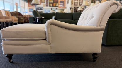 A vintage-style Bassett chaise lounge with a tufted rolled backrest, plush fabric cushion, and turned wooden legs on front casters.