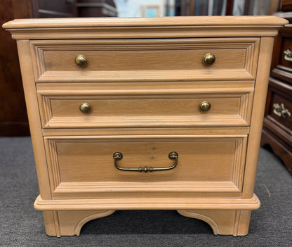 Bernhardt three-drawer wood nightstand with a light finish, routed drawer details, brass knobs, a brass bail pull, and a scalloped base.