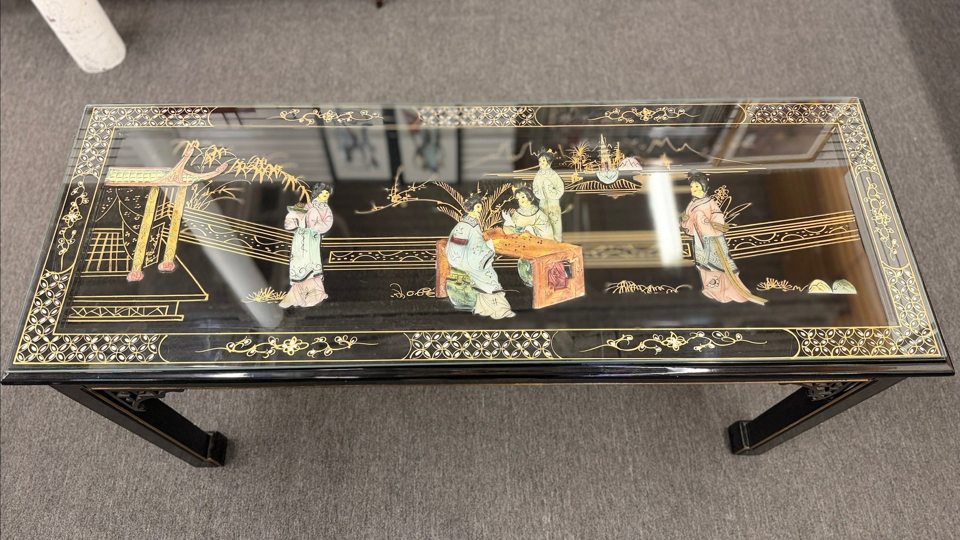 Black lacquer console table featuring hand-painted Asian-inspired scenes with figures, architecture, and landscapes beneath a protective clear glass surface.