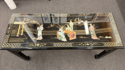 Black lacquer console table featuring hand-painted Asian-inspired scenes with figures, architecture, and landscapes beneath a protective clear glass surface.
