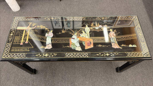 Black lacquer console table featuring hand-painted Asian-inspired scenes with figures, architecture, and landscapes beneath a protective clear glass surface.