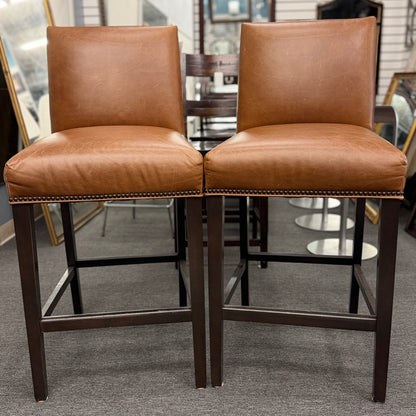 A set of two brown leather barstools featuring nailhead trim details and dark wood legs in good condition.